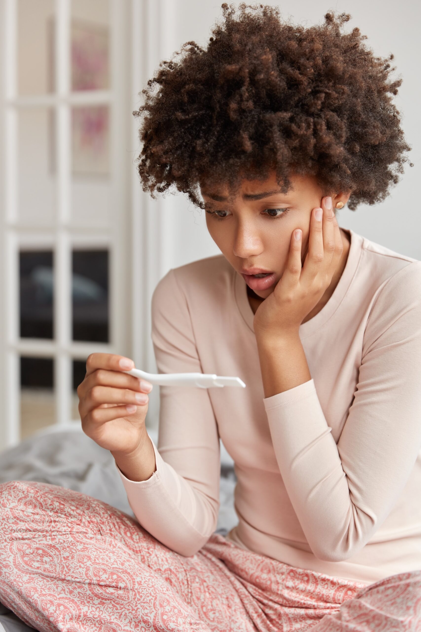 Vertical shot of stupefied woman with Afro haircut, checks positive result on pregnancy test, finds out she expects baby in early age, wears nightclothes, sits at bed. Pregnant shocked girl in bedroom